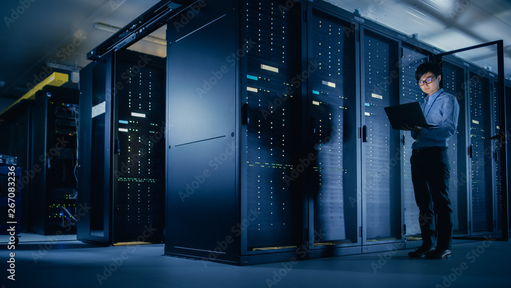 In Data Center: Male IT Technician Running Maintenance Programme on a Laptop, Controls Operational Server Rack Optimal Functioning. Modern High-Tech Telecommunications Operational Super Computer. In Data Center: Male IT Technician Running Maintenance Programme on a Laptop, Controls Operational Server Rack Optimal Functioning. Modern High-Tech Telecommunications Operational Super Computer.
