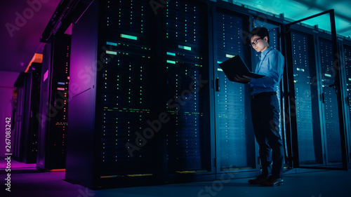 In Data Center: Male IT Technician Running Maintenance Programme on a Laptop, Controls Operational Server Rack Optimal Functioning. Modern High-Tech Operational Super Computer in Neon Colours, Lights photo