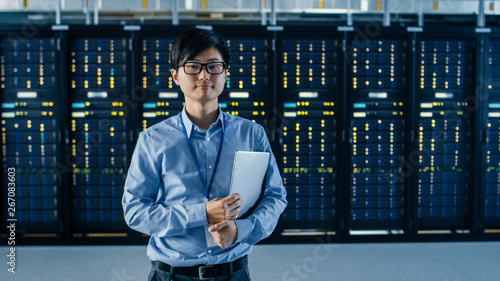 In the Modern Data Center: Portrait of IT Engineer Stands Beside the Row of Server Racks, Finishing Maintenance and Diagnostics Procedure and Holding Laptop.