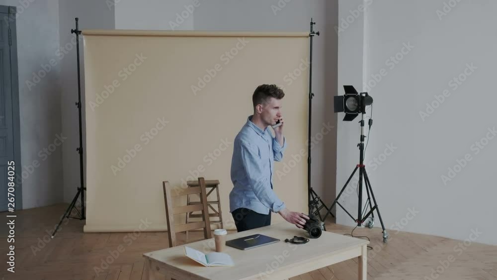 A stylish and busy photographer comes to his workplace in a modern photo studio. He talks on the phone, puts the camera on the table, sits down, opens the laptop and starts working. A stylish and busy photographer comes to his workplace in a modern photo studio. He talks on the phone, puts the camera on the table, sits down, opens the laptop and starts working.