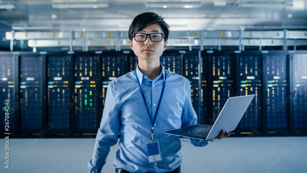 Fototapeta premium In the Modern Data Center: Portrait of IT Engineer Standing with Server Racks Behind Him, Holding Laptop and Looking at the Camera. Finishing Doing Maintenance and Diagnostics Procedure.