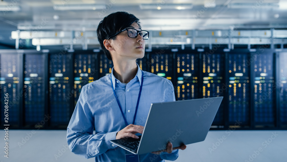 © Gorodenkoff - In the Modern Data Center: Portrait of IT Engineer Stand Near Server Racks, Finishing Maintenance and Diagnostics Procedure Holding Laptop.