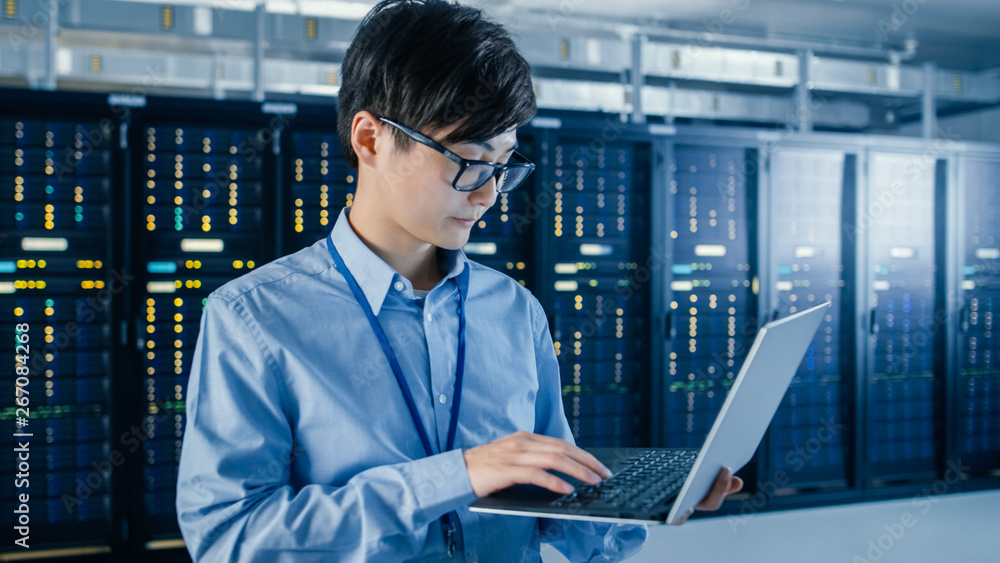In the Modern Data Center: Portrait of IT Engineer Standing with Server Racks Behind Him, Doing Maintenance and Diagnostics Procedure Using Laptop. In the Modern Data Center: Portrait of IT Engineer Standing with Server Racks Behind Him, Doing Maintenance and Diagnostics Procedure Using Laptop.
