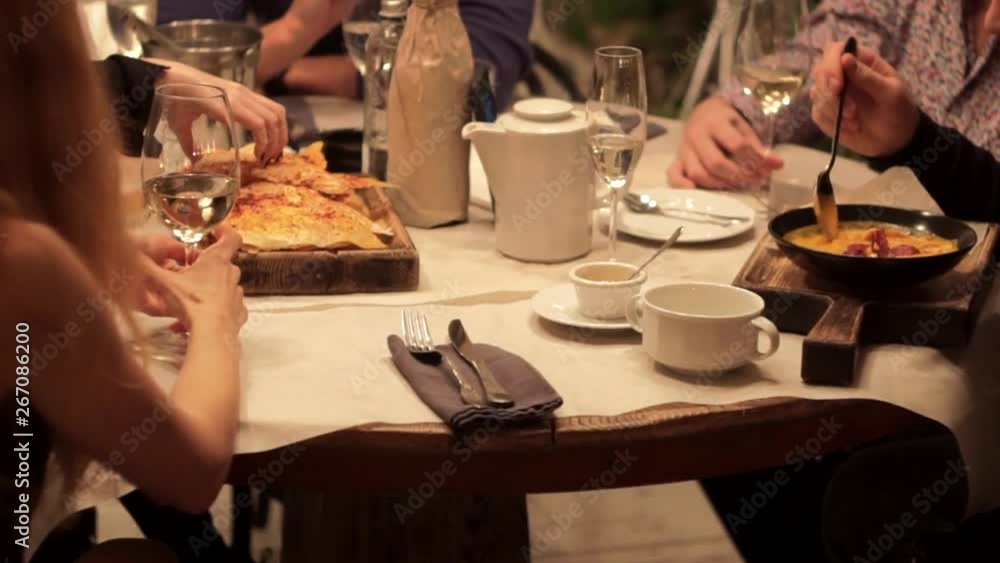 People communicate over evening dinner at a restaurant table under warm muffled lighting. Teapot and cups, wine glasses, cutlery, napkins and dishes on a wooden table covered with white paper. People communicate over evening dinner at a restaurant table under warm muffled lighting. Teapot and cups, wine glasses, cutlery, napkins and dishes on a wooden table covered with white paper.