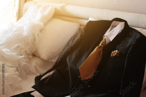 Wedding dress and suit lying on the white bed with beautiful sun light in hotel room. Valentines day and love for celebration concept.