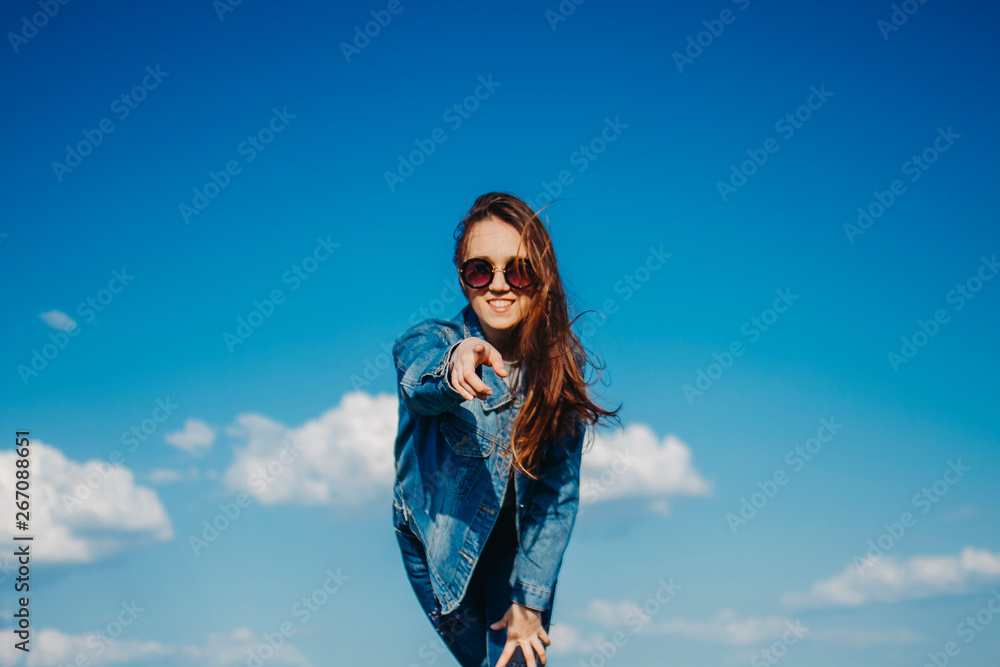 Young woman in sunglasses and denim jacket on blue sky background smiling shows hand to the side Young woman in sunglasses and denim jacket on blue sky background smiling shows hand to the side