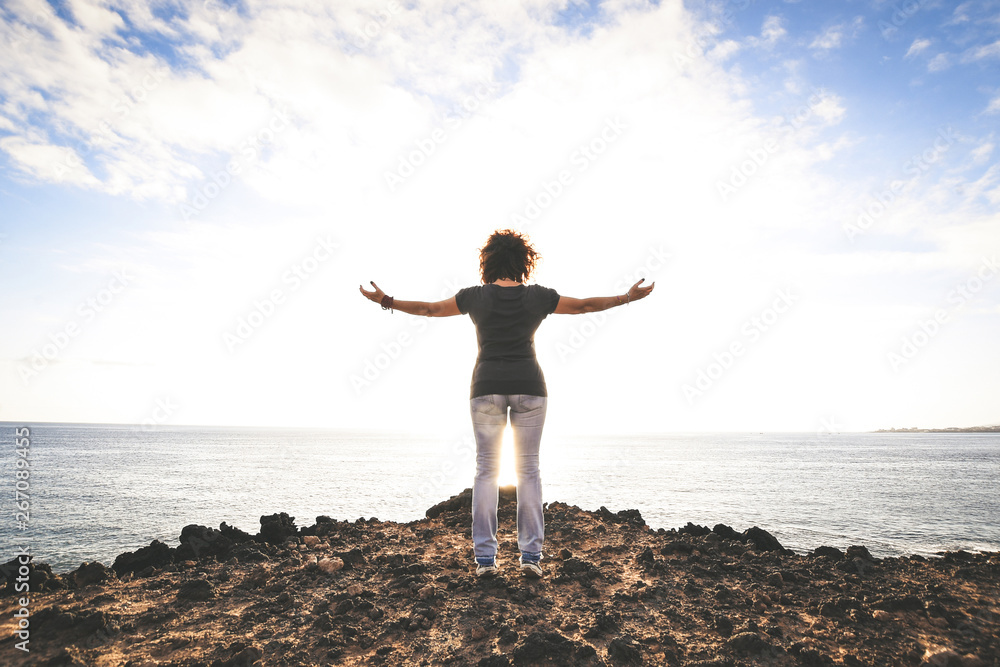 Strong confidence woman with arms open standing on rock at sunset looking ocean.Beautiful girl stay on the peak of mountain cliff edge under sundown light sky enjoying success, freedom, bright future.