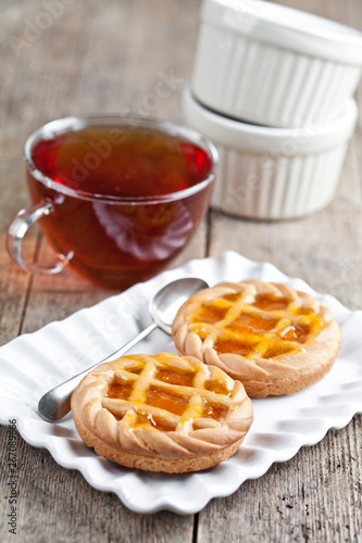 Fresh baked tarts with marmalade or apricot jam filling on white ceramic plate and cup of tea on rustic wooden table. photo