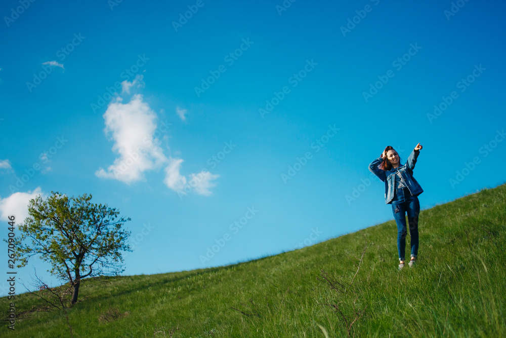 A woman in jeans and a denim jacket stands on top of a hill holding her head from emotion and shows aside