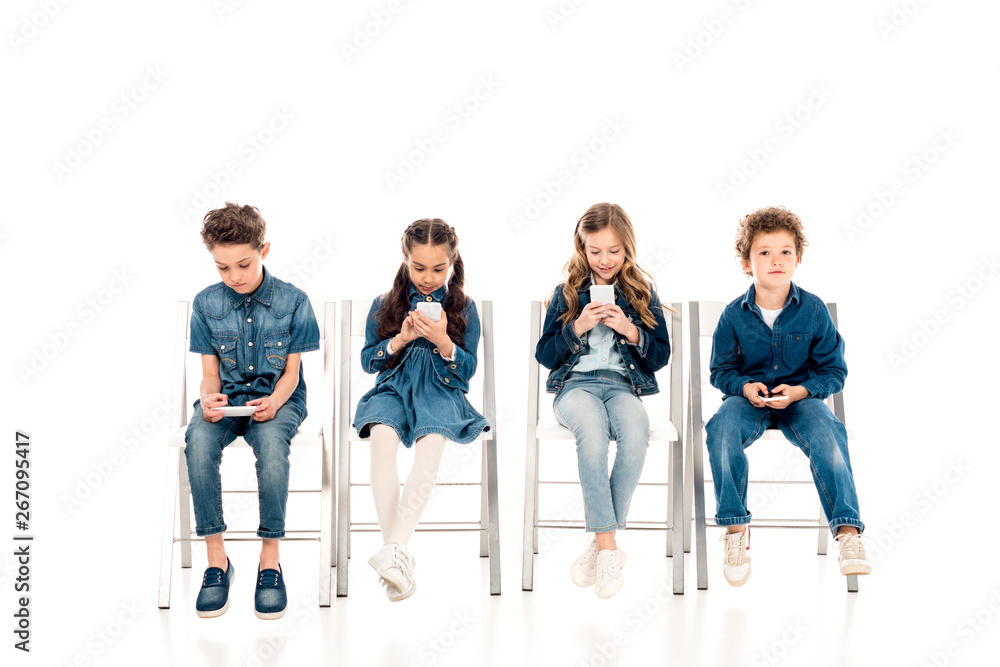 Obraz premium four children in denim clothes sitting on chairs and using smartphones on white