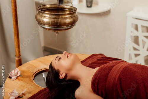 relaxed young woman lying under shirodhara vessel during ayurvedic procedure photo