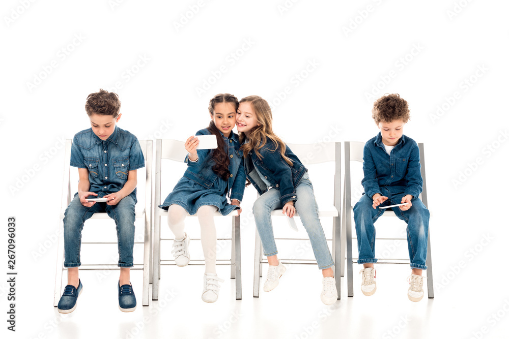 Naklejka premium kids in denim clothes sitting on chairs and taking selfie on white