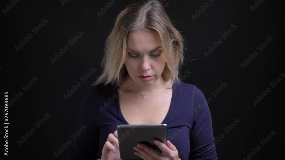 Blonde businesswoman in blue blouse with scoop neckline watching thoughtfully into tablet at black background. Blonde businesswoman in blue blouse with scoop neckline watching thoughtfully into tablet at black background.