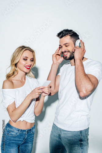 cheerful girl holding smartphone near bearded man in headphones on white