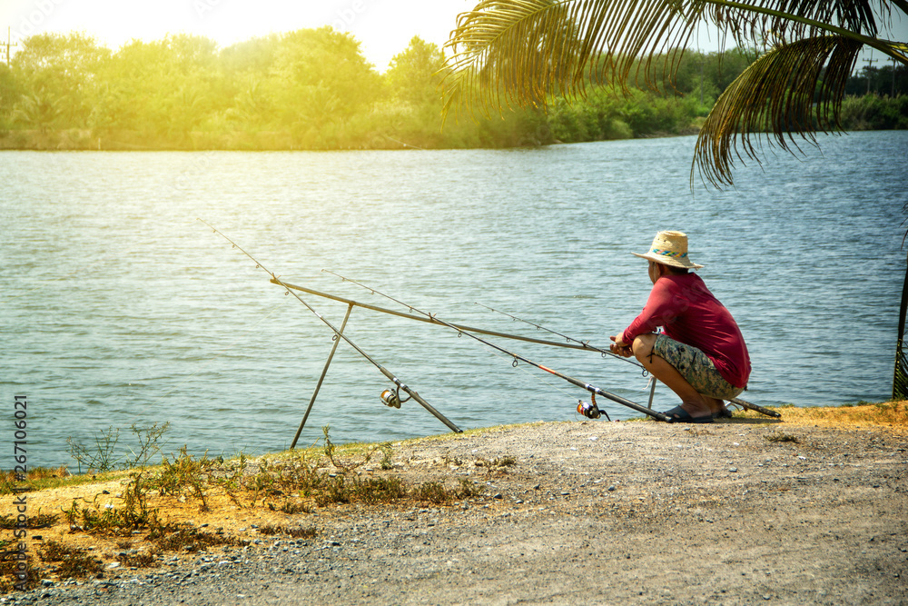 Fishing activities. fisherman middle-aged man red shirt with a straw hat  and three fishing rods on natural water swamp background landscape. Stock  Photo