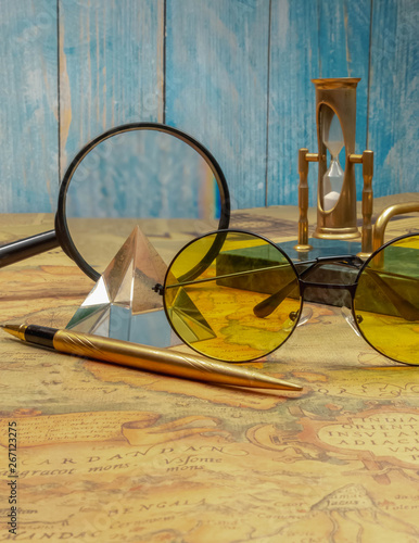 Desktop writing set with an hourglass with a marble base and a crystal pyramid on an ancient map against the background of a travel newspaper and a plank wall the color of sea blue.