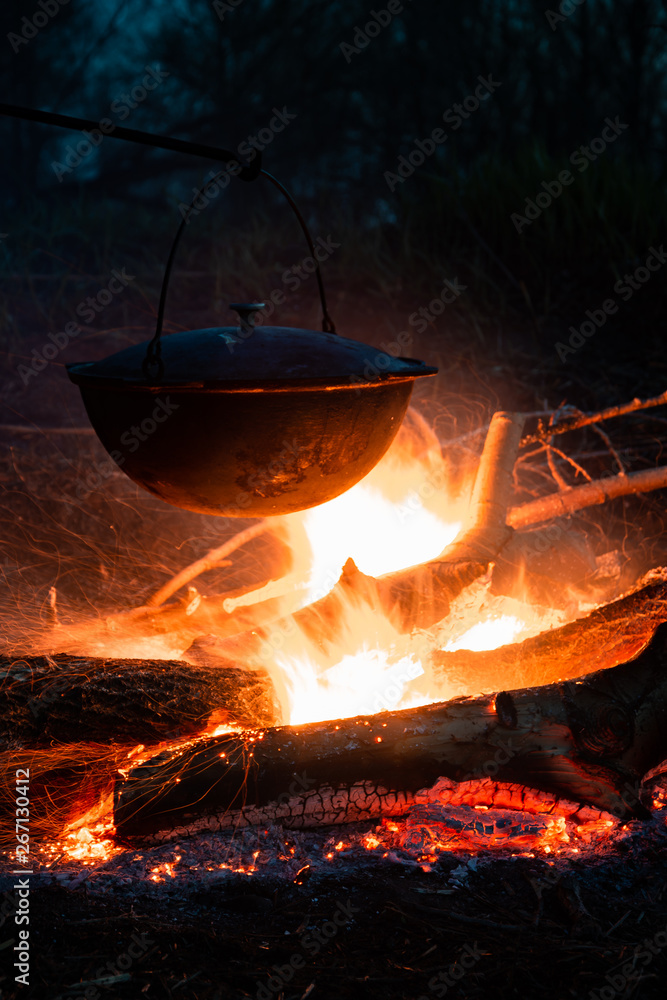 Food cooked on campfire in a large cooking pot, bright flames illuminating the dark scene and metal surface covered with soot, selective focus. Food cooked on campfire in a large cooking pot, bright flames illuminating the dark scene and metal surface covered with soot, selective focus.