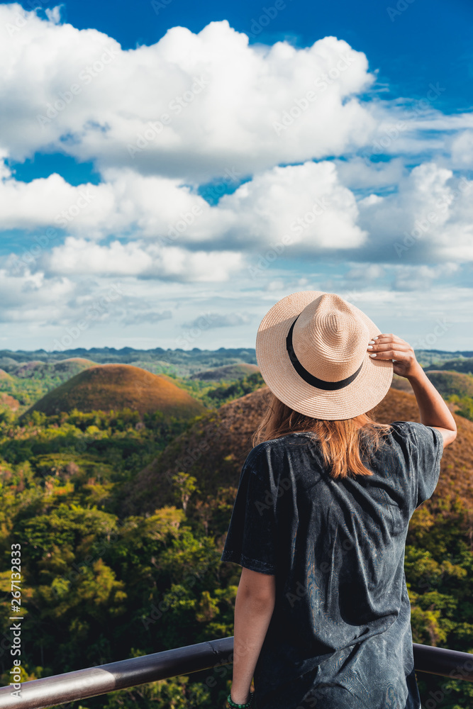 Woman in straw hat standing in front of the Chocolate Hills in the Bohol island in the Philippines. Hills covered in brown grass. Famous touristic place