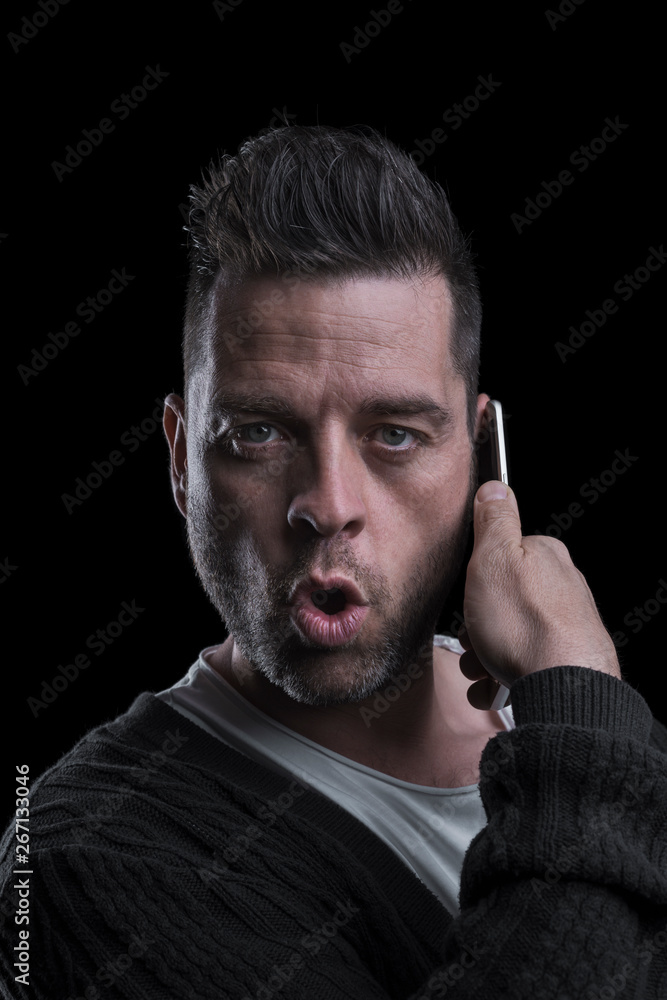 Studio portrait of a surprised man looking at the camera talking on the mobile phone. Isolated on black background. Vertical.