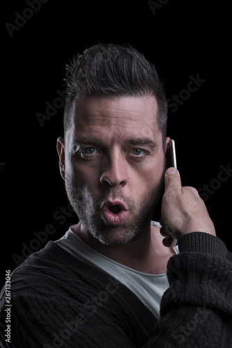Studio portrait of a surprised man looking at the camera talking on the mobile phone. Isolated on black background. Vertical.