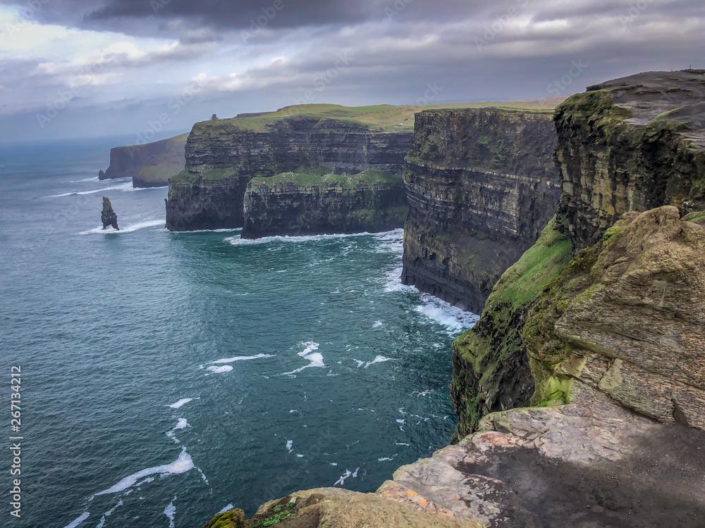 Landscape Shot of the Cliffs of Moher in Ireland - on a Cold Winter Day with Green Grass, Cragged Rocks in the Foreground, Bright Blue water and Cloudy Skies Overhead