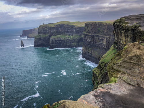 Landscape Shot of the Cliffs of Moher in Ireland - on a Cold Winter Day with Green Grass, Cragged Rocks in the Foreground, Bright Blue water and Cloudy Skies Overhead