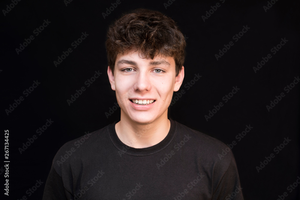 Fototapeta premium A teenager with dark hair in a dark shirt sincerely smiles on a dark background. The boy has a good mood