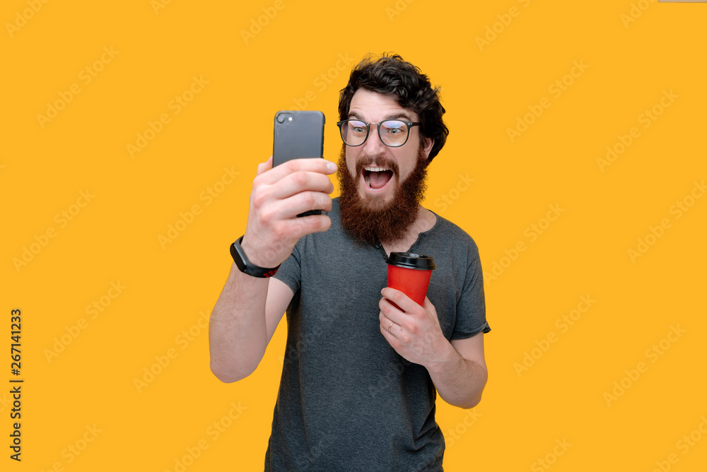 Exited bearded guy holding is taking a photo with smartphone and holding a cup coffee Exited bearded guy holding is taking a photo with smartphone and holding a cup coffee