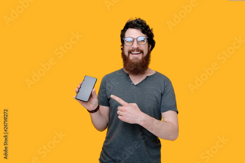 Photo of handsome bearded guy pointing at his mobile while looking at camera