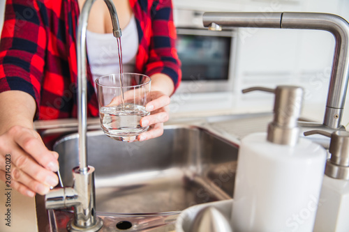 Woman is pouring water from the sink to the glass in the kitchen.