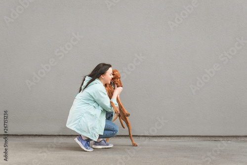 Portrait of a playful dog and a happy woman in casual clothes against a background of gray wall. Owner plays with the dog on the street. Walking with a pet at the weekend