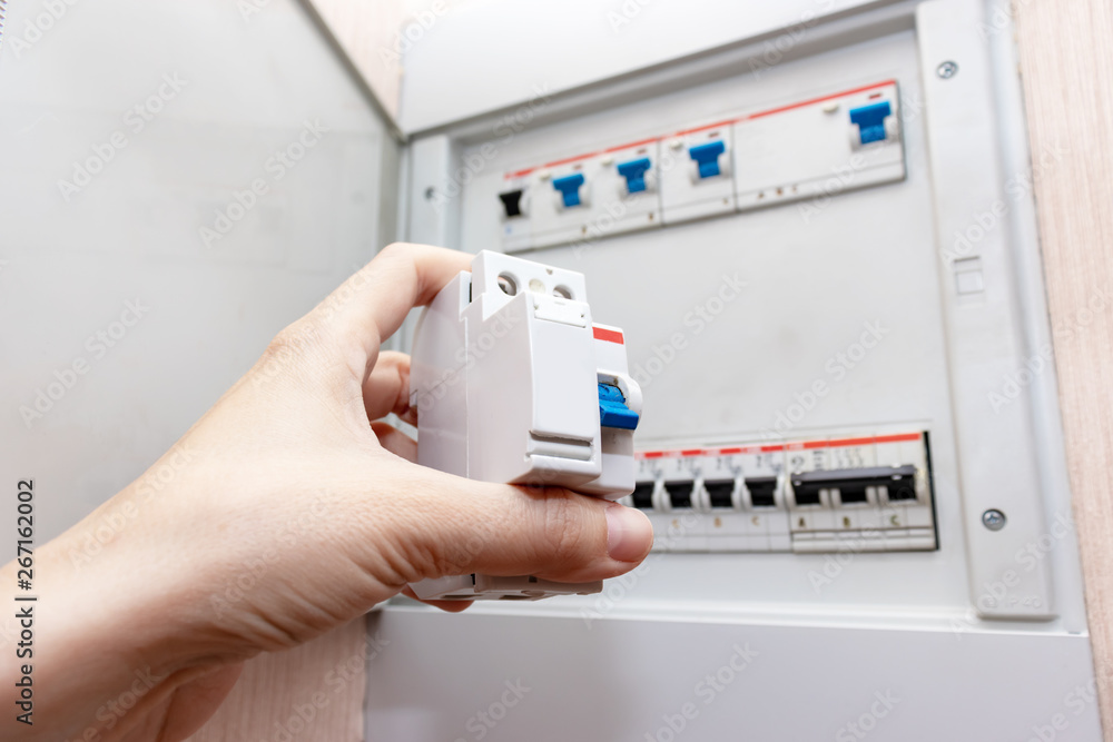 Hand holding new automatic switch and blurred electrical shield with automatic switches of electricity in the house on background- electricity control panel with circuit breakers Hand holding new automatic switch and blurred electrical shield with automatic switches of electricity in the house on background- electricity control panel with circuit breakers