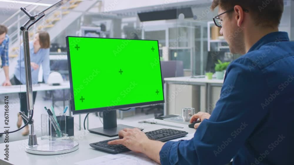 Over the Shoulder Shot of Engineer Working with Green Mock-up Screen Desktop Computer. In the Background Engineering Facility with Specialists Working on Blueprints and Drawings with Industrial Design Over the Shoulder Shot of Engineer Working with Green Mock-up Screen Desktop Computer. In the Background Engineering Facility with Specialists Working on Blueprints and Drawings with Industrial Design