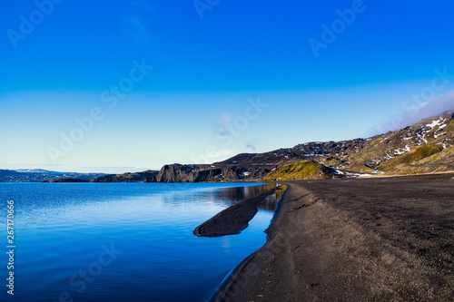 Fototapeta Naklejka Na Ścianę i Meble -  A black beach in Iceland by a clear blue lake, with a mountain chain and a large cliff in the background on a rare summer day