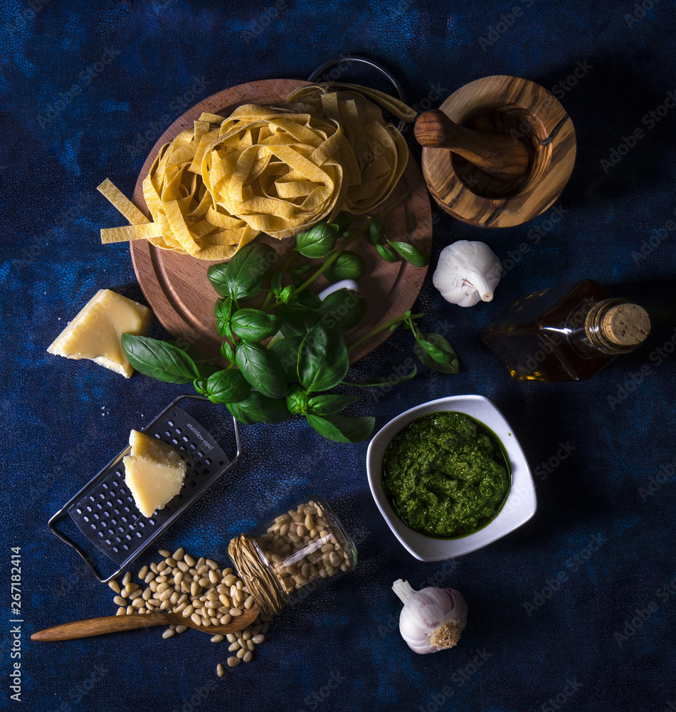 Making pesto. The table with ingredients to produce basil pesto. Mortar, fresh basil herbs, pine nuts, olive oil, parmesan cheese, garlic, bowl with pesto, grater, fresh homemade pasta.