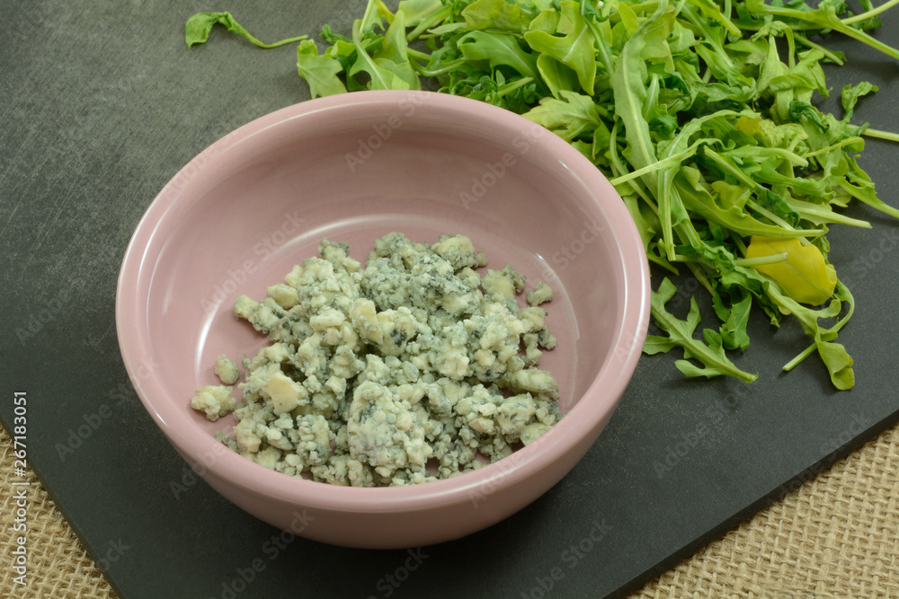 Blue cheese in pink condiment bowl and argula salad leaves on black cutting board on burlap Blue cheese in pink condiment bowl and argula salad leaves on black cutting board on burlap
