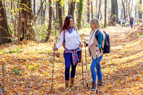 Two young girls walking in forest with backpacks using sticks with fall leaves and trees on background