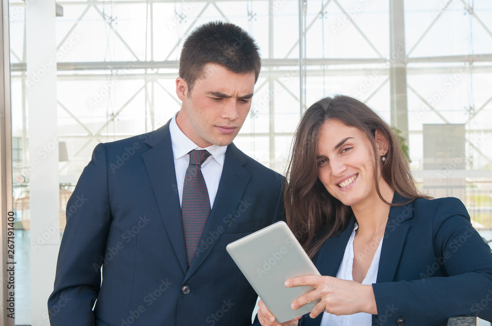 Pretty smiling woman in white blouse and concentrated young man in dark suite standing in office corridor, woman showing data on tablet. Work, communication concept Pretty smiling woman in white blouse and concentrated young man in dark suite standing in office corridor, woman showing data on tablet. Work, communication concept