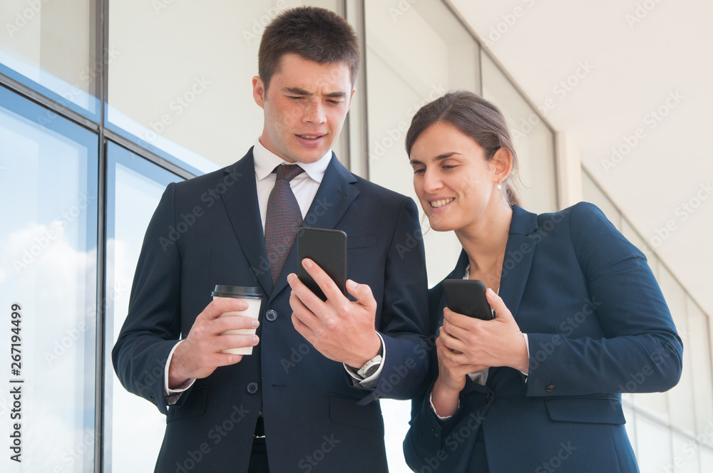 Puzzled manager with takeaway cup showing smartphone screen to female coworker. Two colleagues with phones sharing news during coffee break. Sharing news concept Puzzled manager with takeaway cup showing smartphone screen to female coworker. Two colleagues with phones sharing news during coffee break. Sharing news concept