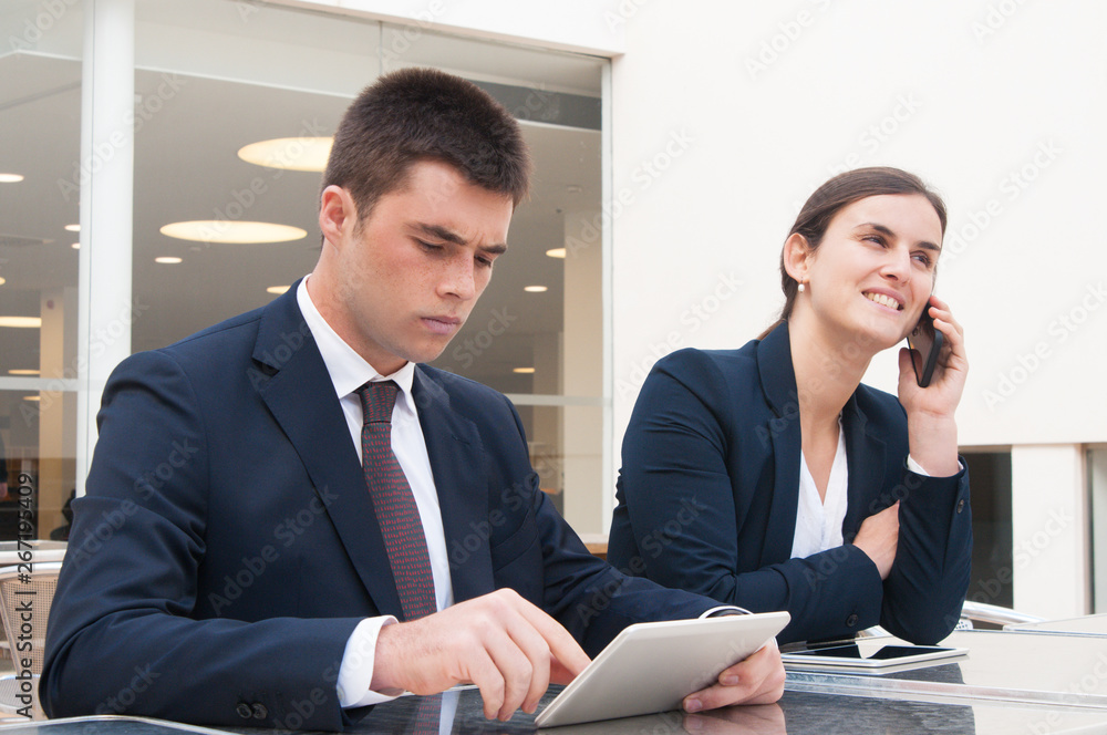 Colleagues using tablet and talking on phone at desk outdoors. Business man and woman wearing formal clothes and sitting at cafe table. Business and communication concept. Colleagues using tablet and talking on phone at desk outdoors. Business man and woman wearing formal clothes and sitting at cafe table. Business and communication concept.