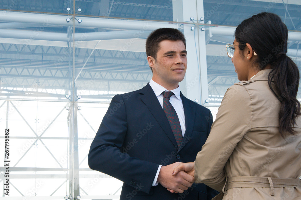 Positive business people meeting and shaking hands outdoors. Business man greeting woman who is standing back to camera with building glass wall in background. Colleagues or partners concept. Positive business people meeting and shaking hands outdoors. Business man greeting woman who is standing back to camera with building glass wall in background. Colleagues or partners concept.