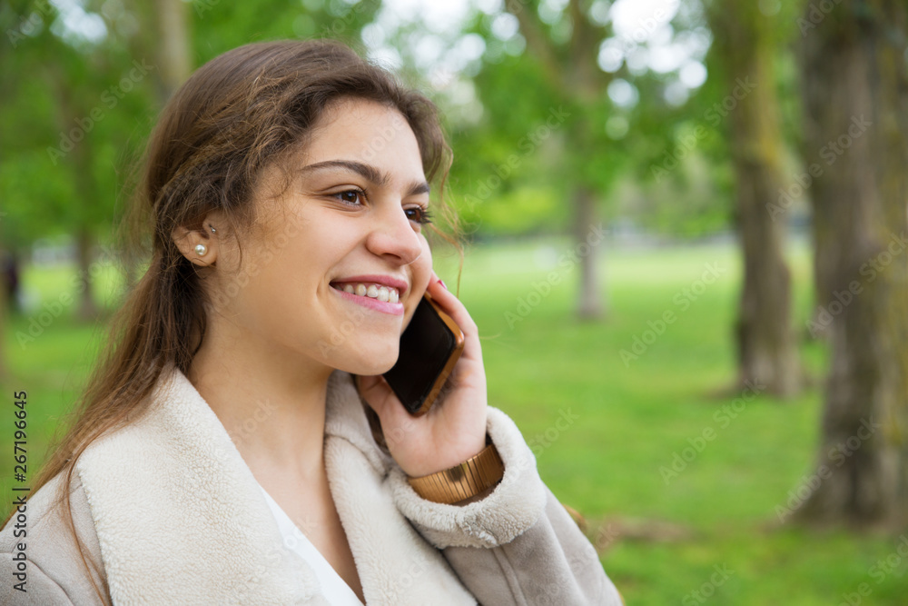 Happy pretty woman calling on smartphone in park. Lady wearing jacket, talking on mobile phone and standing with green grass and trees in background. Communication and nature concept. Happy pretty woman calling on smartphone in park. Lady wearing jacket, talking on mobile phone and standing with green grass and trees in background. Communication and nature concept.