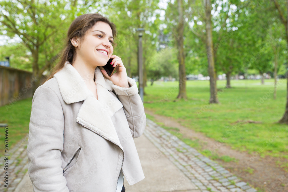 Happy pretty young woman calling on smartphone in park. Lady wearing jacket, talking on mobile phone and standing with walkway and green trees in background. Communication and nature concept. Happy pretty young woman calling on smartphone in park. Lady wearing jacket, talking on mobile phone and standing with walkway and green trees in background. Communication and nature concept.