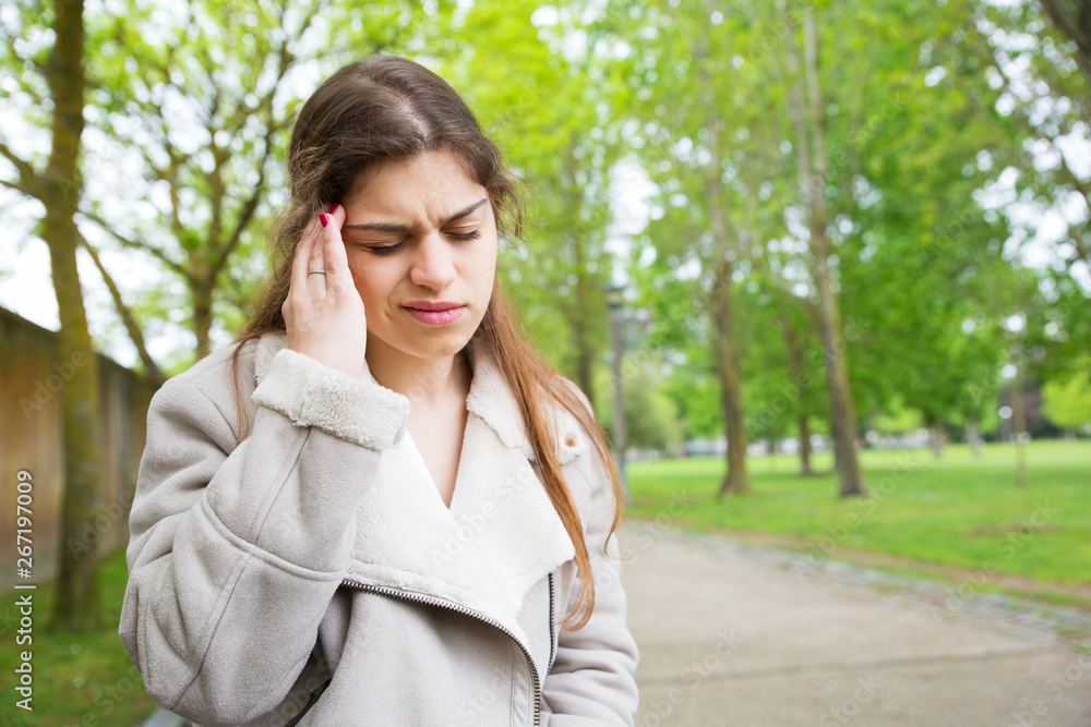 Tired young woman touching temple in park. Lady wearing jacket and standing with closed eyes, walkway and green trees in background. Headache concept. Front view. Tired young woman touching temple in park. Lady wearing jacket and standing with closed eyes, walkway and green trees in background. Headache concept. Front view.