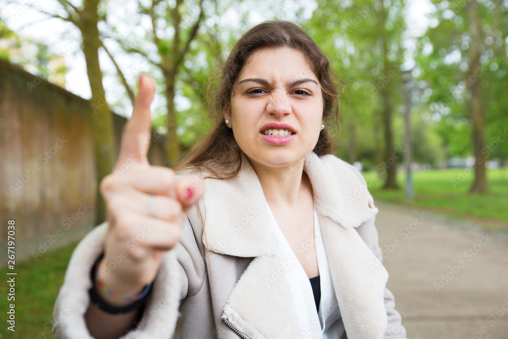 Angry nervous girl threatening at camera. Young woman in casual jacket standing in park and waging finger. Threatening concept Angry nervous girl threatening at camera. Young woman in casual jacket standing in park and waging finger. Threatening concept