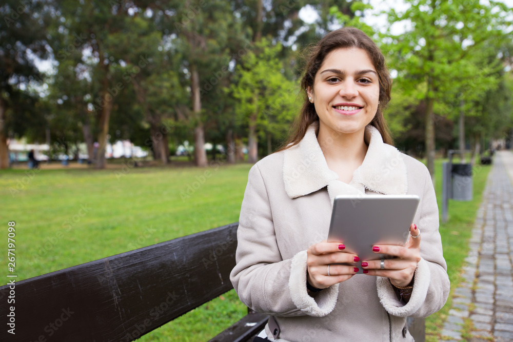 Cheerful Latin girl relaxing in park. Young woman in warm jacket sitting on bench and using tablet, grass and trees in background. Communication concept Cheerful Latin girl relaxing in park. Young woman in warm jacket sitting on bench and using tablet, grass and trees in background. Communication concept