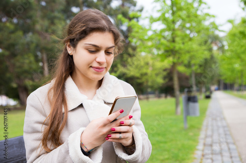 Positive peaceful girl texting message on mobile phone. Young woman in warm casual jacket sitting on bench in park and using phone. Connection outdoors concept