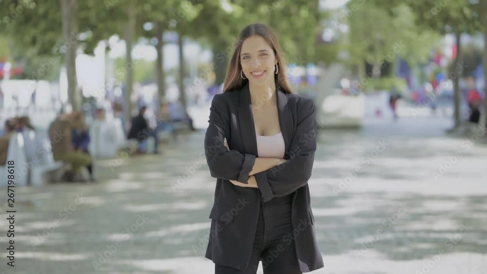 Attractive young woman smiling at camera outdoor. Front view of beautiful happy girl standing with crossed arms and hand in waist on street. Beauty concept