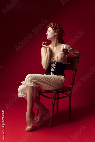 Medieval redhead young woman as a duchess in black corset and night clothes sitting on a chair on red background with a glass of wine. Concept of comparison of eras, modernity and renaissance.