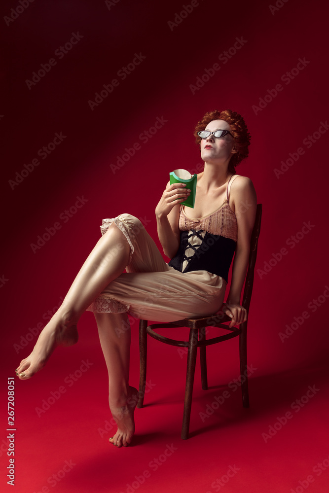 Medieval redhead young woman as a duchess in black corset, sunglasses and night clothes sitting on red background eating fried potato. Concept of comparison of eras, modernity and renaissance. Medieval redhead young woman as a duchess in black corset, sunglasses and night clothes sitting on red background eating fried potato. Concept of comparison of eras, modernity and renaissance.